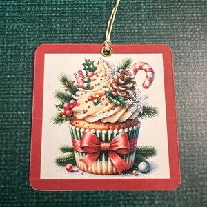 May include: A red and white Christmas cupcake ornament with a green and white striped paper liner, a red bow, and a candy cane. The cupcake is decorated with white frosting, green holly leaves, red berries, and a pine cone.