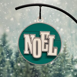 May include: A round, teal Christmas ornament with the word "NOEL" in white, outlined in gray. The ornament hangs from a black metal hook against a snowy, evergreen forest backdrop. A festive holiday decoration.