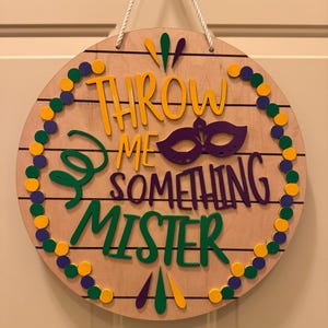 May include: A round wooden sign with the text "THROW ME SOMETHING MISTER" in yellow, green, and purple. A purple mask graphic is in the center. The sign is decorated with yellow, green, and purple beads around the edge and has a white rope for hanging.