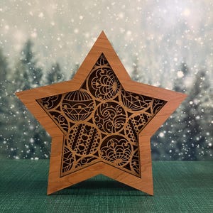 Pode incluir: Decoração em forma de estrela de madeira com intrincados desenhos recortados. A estrela, de madeira castanha clara, apresenta um padrão detalhado de formas circulares e geométricas. O fundo mostra uma cena de floresta nevada, evocando o inverno.