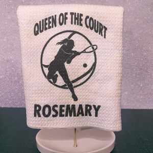 Pode incluir: Toalha branca com texto preto que diz "Queen of the Court" e uma silhueta de uma jogadora de tênis segurando uma raquete. O nome "Rosemary" está impresso abaixo da imagem.