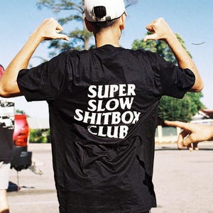May include: A black t-shirt with the text "SUPER SLOW SHITBOX CLUB" in white. The person is wearing black shorts and a white baseball cap. Another person is visible in the background wearing a black t-shirt.
