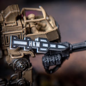 Op de afbeelding: Een close-up van een speelgoed militaire voertuig met een zwart-witte raket bevestigd aan de zijkant. De raket heeft de letters "HKM" erop gedrukt.