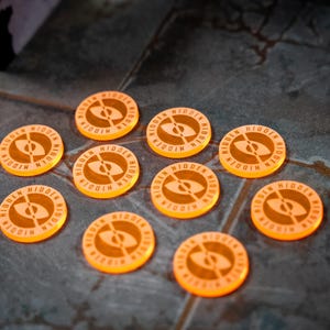 Op de afbeelding: Tien oranje spel fiches met het woord "HIDDEN" en een oog symbool. De fiches liggen verspreid op een donker, getextureerd oppervlak, wat wijst op een bordspel.