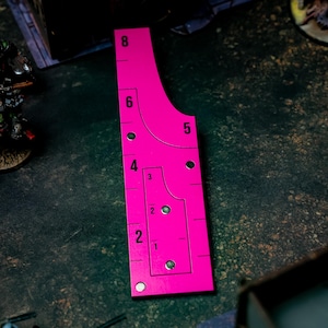 May include: A bright pink gaming range ruler with measurements from 1 to 8 inches. The ruler has curved edges and circular cutouts. It is designed for tabletop games and is placed on a dark, textured surface.