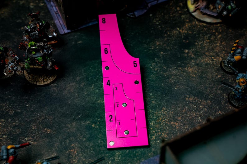May include: A bright pink gaming range ruler with measurements from 1 to 8 inches. The ruler has curved edges and circular cutouts. It is designed for tabletop games and is placed on a dark, textured surface.