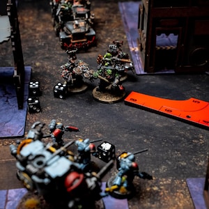 May include: A tabletop war game scene featuring miniature figures, dice, and terrain. The figures are painted in various colors and are positioned on a textured surface. A red measuring tool is also present.