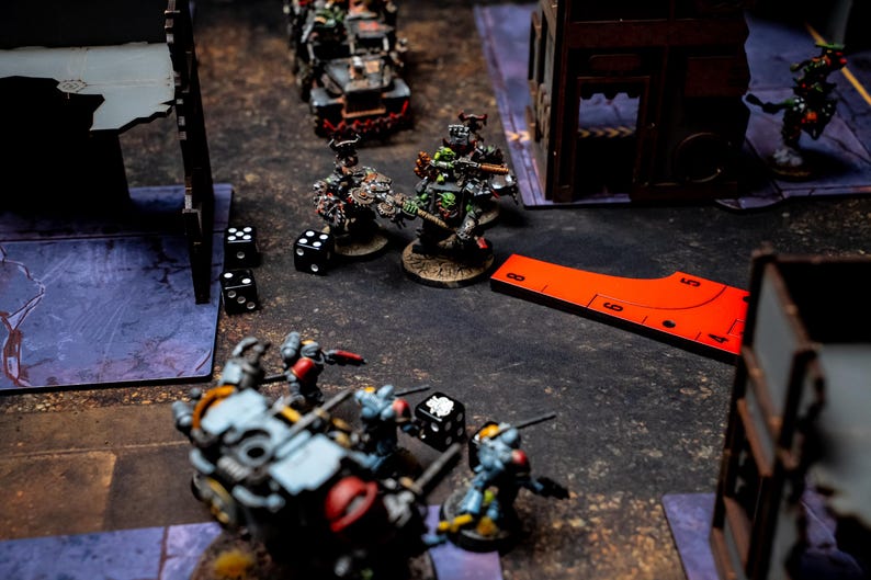May include: A tabletop war game scene featuring miniature figures, dice, and terrain. The figures are painted in various colors and are positioned on a textured surface. A red measuring tool is also present.