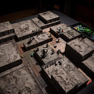 K&ouml;nnte beinhalten: Ein detailliertes Tabletop-Rollenspiel-Gel&auml;nde. Die Szene zeigt modulare, strukturierte graue Strukturen, die angeordnet sind, um ein Schlachtfeld darzustellen. Miniaturfiguren und W&uuml;rfel sind &uuml;ber die Landschaft verstreut.
