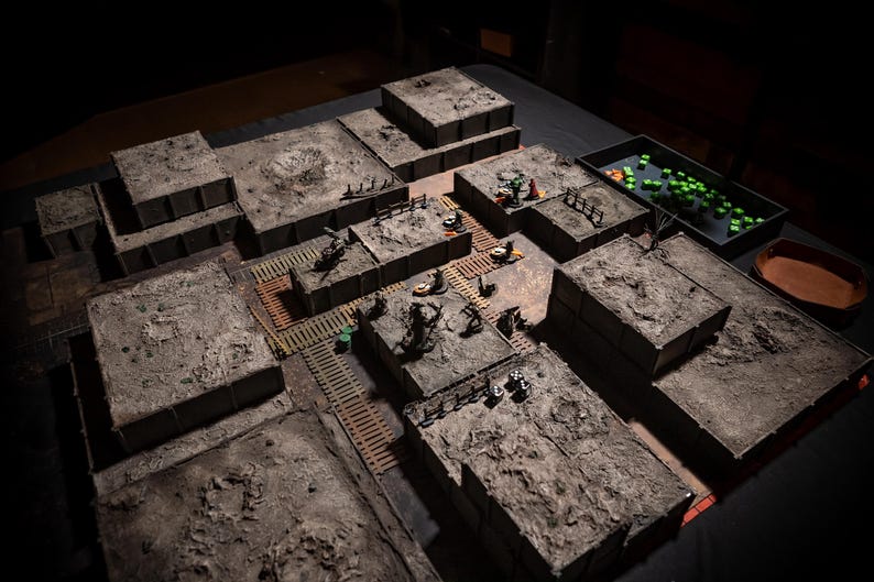 K&ouml;nnte beinhalten: Ein detailliertes Tabletop-Rollenspiel-Gel&auml;nde. Die Szene zeigt modulare, strukturierte graue Strukturen, die angeordnet sind, um ein Schlachtfeld darzustellen. Miniaturfiguren und W&uuml;rfel sind &uuml;ber die Landschaft verstreut.