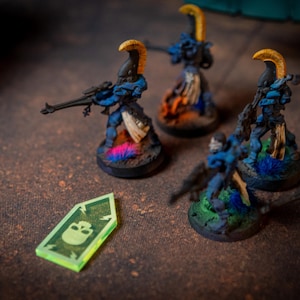 Puede incluir: Primer plano de un conjunto de figuras en miniatura azules y doradas con un símbolo de calavera verde en una ficha de plástico transparente. Las figuras están de pie sobre bases redondas con pintura azul y verde.