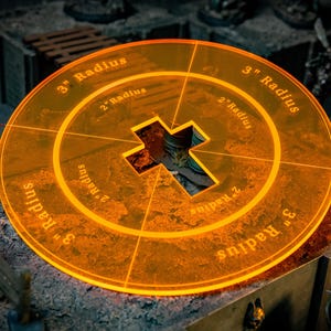 Pode incluir: Um acessório de jogo circular laranja transparente, com marcações de raio de 5 cm e 7,6 cm. O centro apresenta um recorte em forma de cruz. O texto "3" Radius" e "2" Radius" é visível. Ideal para jogos de tabuleiro.