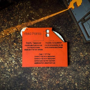 Op de afbeelding: Een rode spelkaart met de tekst "Yield Points" en spelinstructies. De kaart heeft een witte rand en is gedeeltelijk gescheurd. De kaart ligt op een getextureerd, bruin oppervlak.