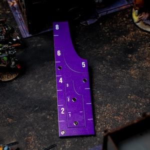 May include: A purple gaming range ruler with white numerical markings from 1 to 8. The ruler has a curved edge and several holes. Miniature figures are visible in the background, suggesting a tabletop game setting.