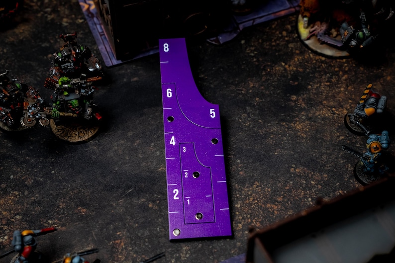 May include: A purple gaming range ruler with white numerical markings from 1 to 8. The ruler has a curved edge and several holes. Miniature figures are visible in the background, suggesting a tabletop game setting.