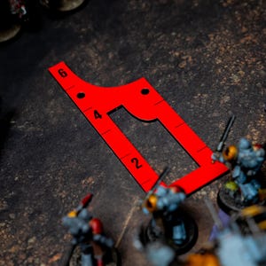 May include: A bright red, angular game ruler with measurements from 2 to 6, used in tabletop gaming. The ruler is on a textured, dark surface, with miniature figures and game elements in the background.
