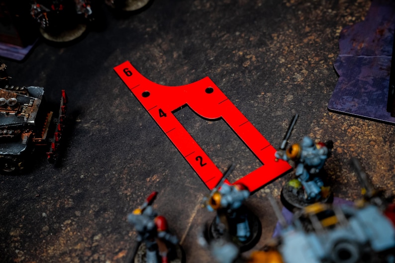 May include: A bright red, angular game ruler with measurements from 2 to 6, used in tabletop gaming. The ruler is on a textured, dark surface, with miniature figures and game elements in the background.