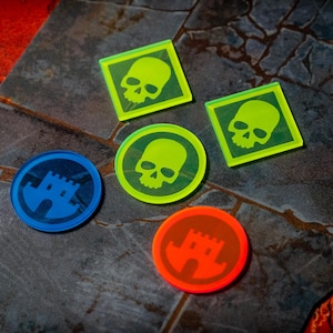 Puede incluir: Una colección de cinco fichas de juego de acrílico translúcido. Dos fichas cuadradas de color verde neón presentan un icono de calavera. Una ficha circular de color verde neón presenta un icono de calavera. Una ficha azul y una naranja presentan un castillo.