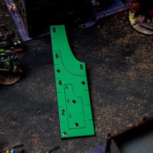 May include: A bright green gaming template with black markings and numbers, used for measuring distances in tabletop games. The template has curved and straight edges, with numbered increments from 1 to 8 inches. Miniature figures are visible in the background.