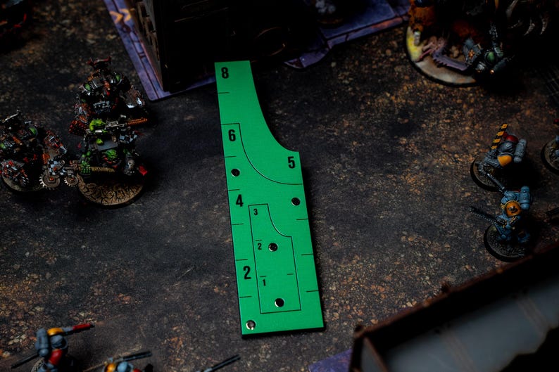 May include: A bright green gaming template with black markings and numbers, used for measuring distances in tabletop games. The template has curved and straight edges, with numbered increments from 1 to 8 inches. Miniature figures are visible in the background.