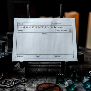 May include: A blank white card with headings for "Name", "Type", "Stats", "Rarity", "Cost", "Bulk", "Campaign", "Notes", and "Attunement". The card is on a wooden easel and there are dice and coins scattered around it.
