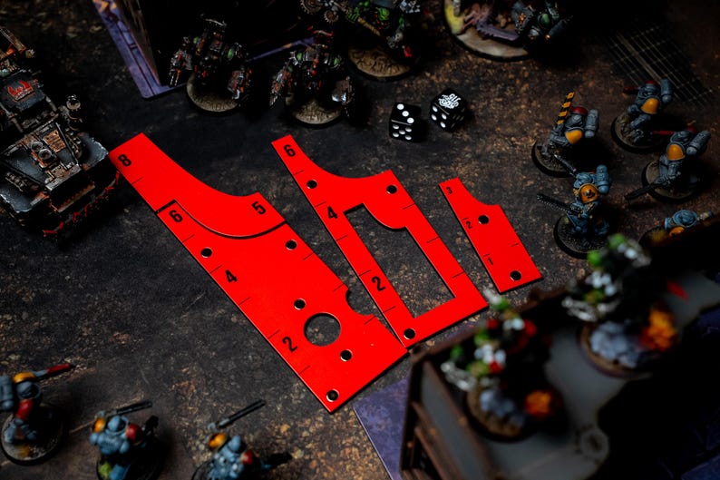 May include: Bright red gaming templates with measurement markings, including a curved ruler and two other shapes. The templates are on a dark surface with miniature figurines and dice in the background. The templates are used for tabletop games.