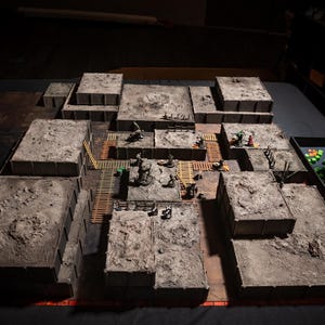 K&ouml;nnte beinhalten: Ein detailliertes Tabletop-Gaming-Gel&auml;nde mit mehreren grauen, strukturierten Plattformen, die wie eine Stadt oder Festung angeordnet sind. Miniaturfiguren sind auf den Plattformen positioniert, mit Holzstegen und Z&auml;unen. Eine Schale mit gr&uuml;nen W&uuml;rfeln ist sichtbar.