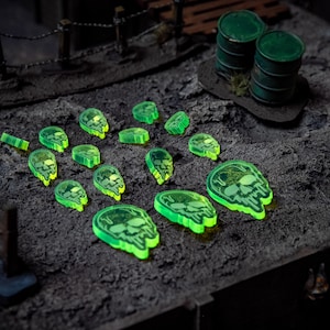May include: A collection of glowing green skull-shaped game tokens and rectangular markers are scattered on a textured gray surface. Two green barrels and a wooden fence are in the background. The tokens have a skull design.