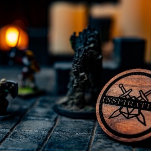 May include: A wooden coaster with a black and white design of two crossed swords and a dice with the word "Inspiration" written on it.