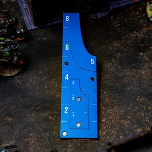 May include: A blue measuring tool with white markings for tabletop gaming. The tool has curved edges and numbered increments from 1 to 8 inches. It is placed on a textured surface with miniature figures in the background.