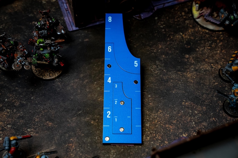 May include: A blue measuring tool with white markings for tabletop gaming. The tool has curved edges and numbered increments from 1 to 8 inches. It is placed on a textured surface with miniature figures in the background.