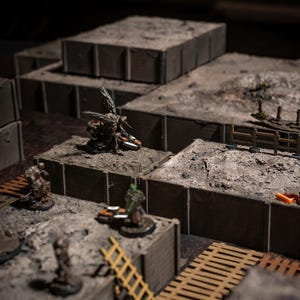 K&ouml;nnte beinhalten: Eine Miniatur-Tabletop-Kriegsspielszene mit mehreren Gel&auml;ndeteilen. Die Szene umfasst modulare, graue, blockartige Strukturen mit sandigen Oberfl&auml;chen, Miniaturfiguren, Leitern und Holzstegen. Die Beleuchtung ist ged&auml;mpft und erzeugt einen dramatischen Effekt.