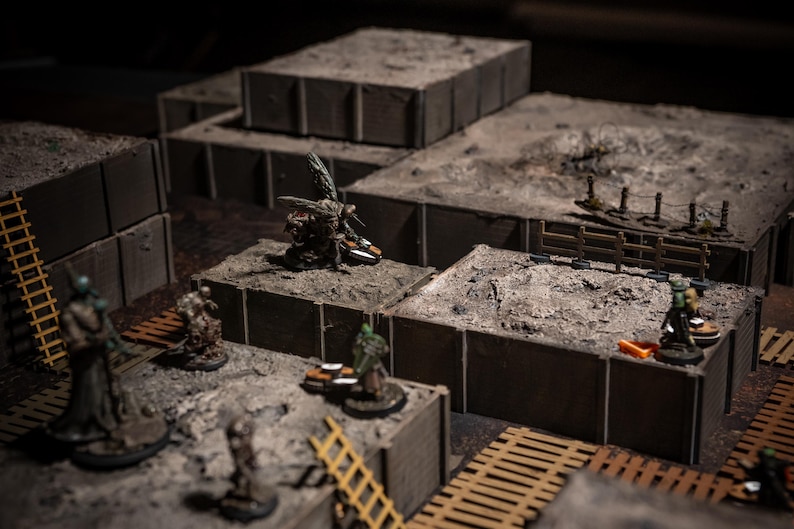 K&ouml;nnte beinhalten: Eine Miniatur-Tabletop-Kriegsspielszene mit mehreren Gel&auml;ndeteilen. Die Szene umfasst modulare, graue, blockartige Strukturen mit sandigen Oberfl&auml;chen, Miniaturfiguren, Leitern und Holzstegen. Die Beleuchtung ist ged&auml;mpft und erzeugt einen dramatischen Effekt.