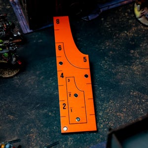 May include: An orange measuring tool with black markings and numbers, used for tabletop gaming. The tool has a curved edge and a series of holes and lines for precise measurements. Numbers range from 1 to 8 inches.