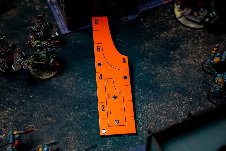 May include: An orange measuring tool with black markings and numbers, used for tabletop gaming. The tool has a curved edge and a series of holes and lines for precise measurements. Numbers range from 1 to 8 inches.