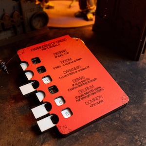 May include: A red game card with white tabs and black text. The card has the text "HARBINGERS OF DREAD" and various game effects listed. The numbers 3, 5, and 6 are visible on the card. The card is on a dark surface.