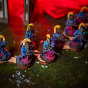 Puede incluir: Una fila de figuras en miniatura azules y amarillas con pistolas, de pie sobre bases circulares. Las figuras est&aacute;n dispuestas en l&iacute;nea sobre una superficie verde.