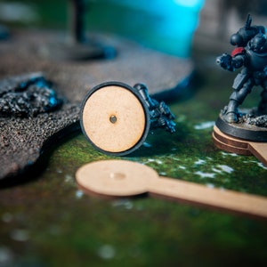 Puede incluir: Una figura de Space Marine en miniatura pintada en negro y amarillo con una hombrera roja, de pie sobre una base redonda de madera. La figura sostiene un arma y est&aacute; en una postura de combate. La figura est&aacute; sobre una superficie texturizada verde y marr&oacute;n.