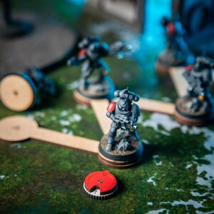 Puede incluir: Una escena de juego de guerra en miniatura con cinco figuras pintadas de gris y rojo, cada una sobre una base circular de madera. Las figuras est&aacute;n conectadas por palos de madera, formando un camino. Un token circular rojo y negro est&aacute; en primer plano.
