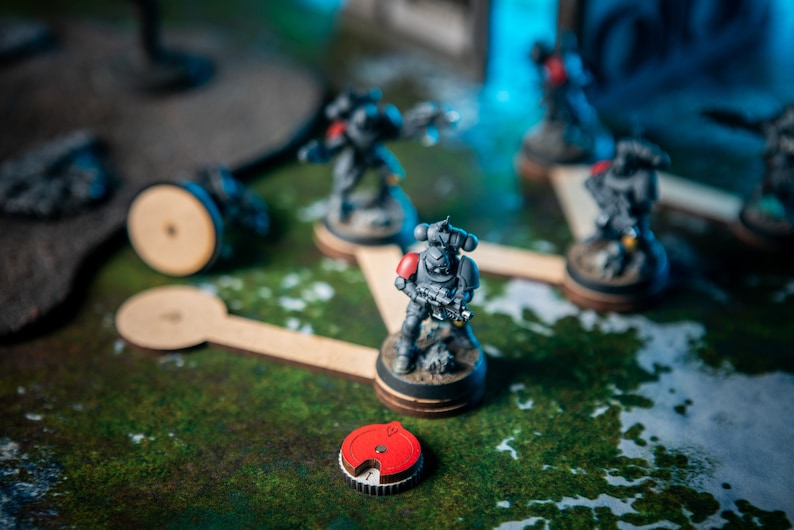 Puede incluir: Una escena de juego de guerra en miniatura con cinco figuras pintadas de gris y rojo, cada una sobre una base circular de madera. Las figuras est&aacute;n conectadas por palos de madera, formando un camino. Un token circular rojo y negro est&aacute; en primer plano.