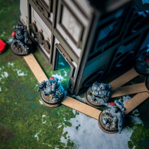 Puede incluir: Un grupo de figuras en miniatura grises y negras, que representan soldados, est&aacute;n colocadas en una estera de juego verde y blanca. Las figuras est&aacute;n de pie sobre bases redondas, y algunas est&aacute;n conectadas por palos de madera. Una estructura de edificio de madera est&aacute; en el fondo.