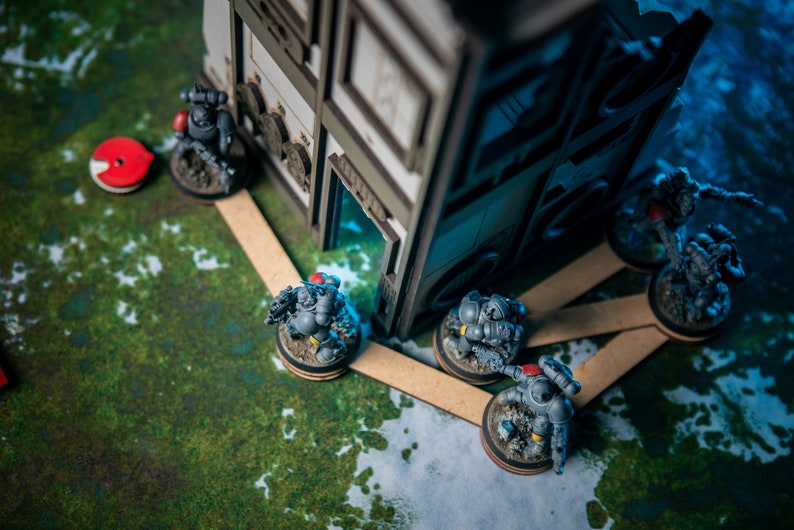 Puede incluir: Un grupo de figuras en miniatura grises y negras, que representan soldados, est&aacute;n colocadas en una estera de juego verde y blanca. Las figuras est&aacute;n de pie sobre bases redondas, y algunas est&aacute;n conectadas por palos de madera. Una estructura de edificio de madera est&aacute; en el fondo.