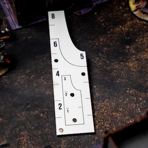 May include: A white, rectangular game measuring tool with black markings and numbers from 1 to 8. The tool has curved and straight edges, with circular cutouts. Miniature figurines are visible in the background, suggesting a tabletop game.