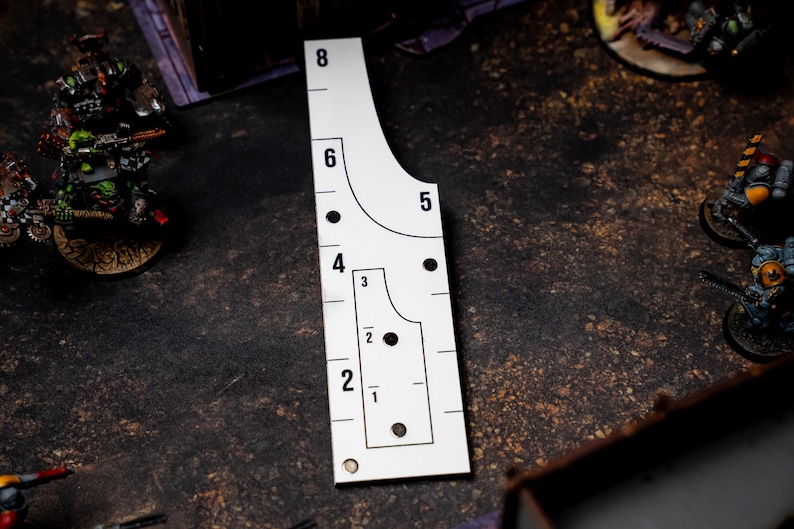 May include: A white, rectangular game measuring tool with black markings and numbers from 1 to 8. The tool has curved and straight edges, with circular cutouts. Miniature figurines are visible in the background, suggesting a tabletop game.