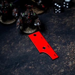 May include: A red measuring tool with black markings and numbers, used in tabletop gaming. The tool has a curved edge and two holes. Miniature figures and dice are in the background, on a dark, textured surface.