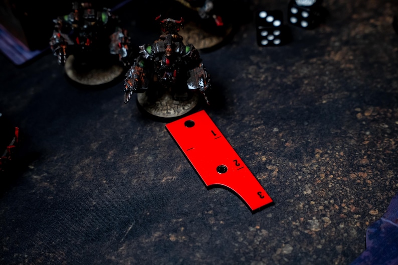 May include: A red measuring tool with black markings and numbers, used in tabletop gaming. The tool has a curved edge and two holes. Miniature figures and dice are in the background, on a dark, textured surface.