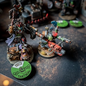 Pode incluir: Duas figuras em miniatura de orcs pintadas de verde e vermelho, uma segurando uma arma grande e a outra montando um veículo com uma arma, sobre uma superfície marrom com fichas circulares verdes e brancas que dizem "Shoota Drills" e "Taktikal".