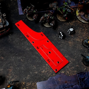 May include: A red game ruler with measurements and cutouts, next to dice and miniature figures. The ruler has markings for distances, with numbers from 2 to 8. The scene suggests a tabletop war game.