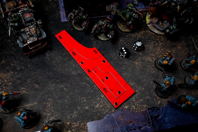 May include: A red game ruler with measurements and cutouts, next to dice and miniature figures. The ruler has markings for distances, with numbers from 2 to 8. The scene suggests a tabletop war game.