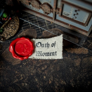 Puede incluir: Primer plano de una pequeña pancarta de tela blanca con el texto "Oath of Moment" impreso en negro. Un sello de cera roja con un diseño de calavera está unido a la pancarta.